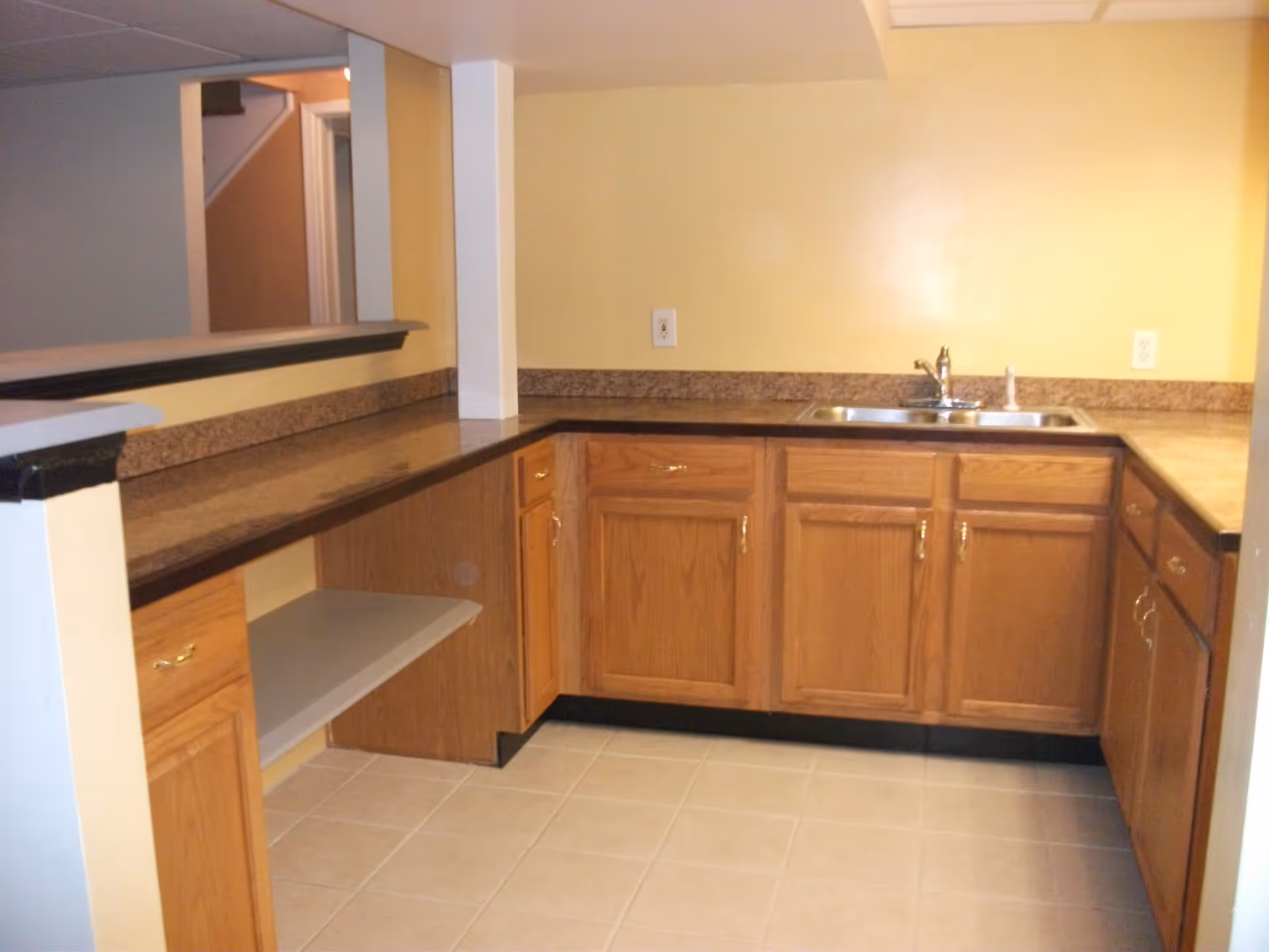 Interior view of a kitchen area with wooden cabinets, a double sink, and a countertop with a brown speckled surface. The walls are painted yellow and the floor is tiled in a light color. There is an open shelf under the counter and a pass-through window opening to another room.