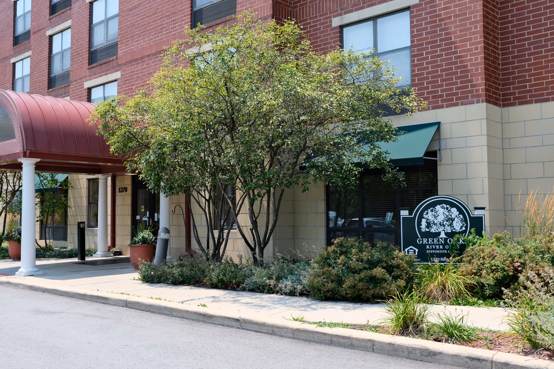 Exterior view of the entrance to Green Oaks of River Oaks facility, showing a red canopy over the doorway, a tree and bushes in front, and a green sign with the facility's name and logo.