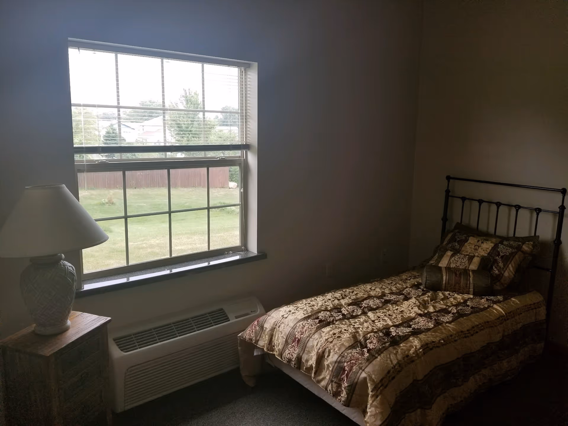 A small bedroom with a single bed featuring a decorative patterned bedspread and pillows. Next to the bed is a wooden nightstand with a lamp. A large window with blinds lets in natural light and shows a grassy outdoor area with trees and a fence.