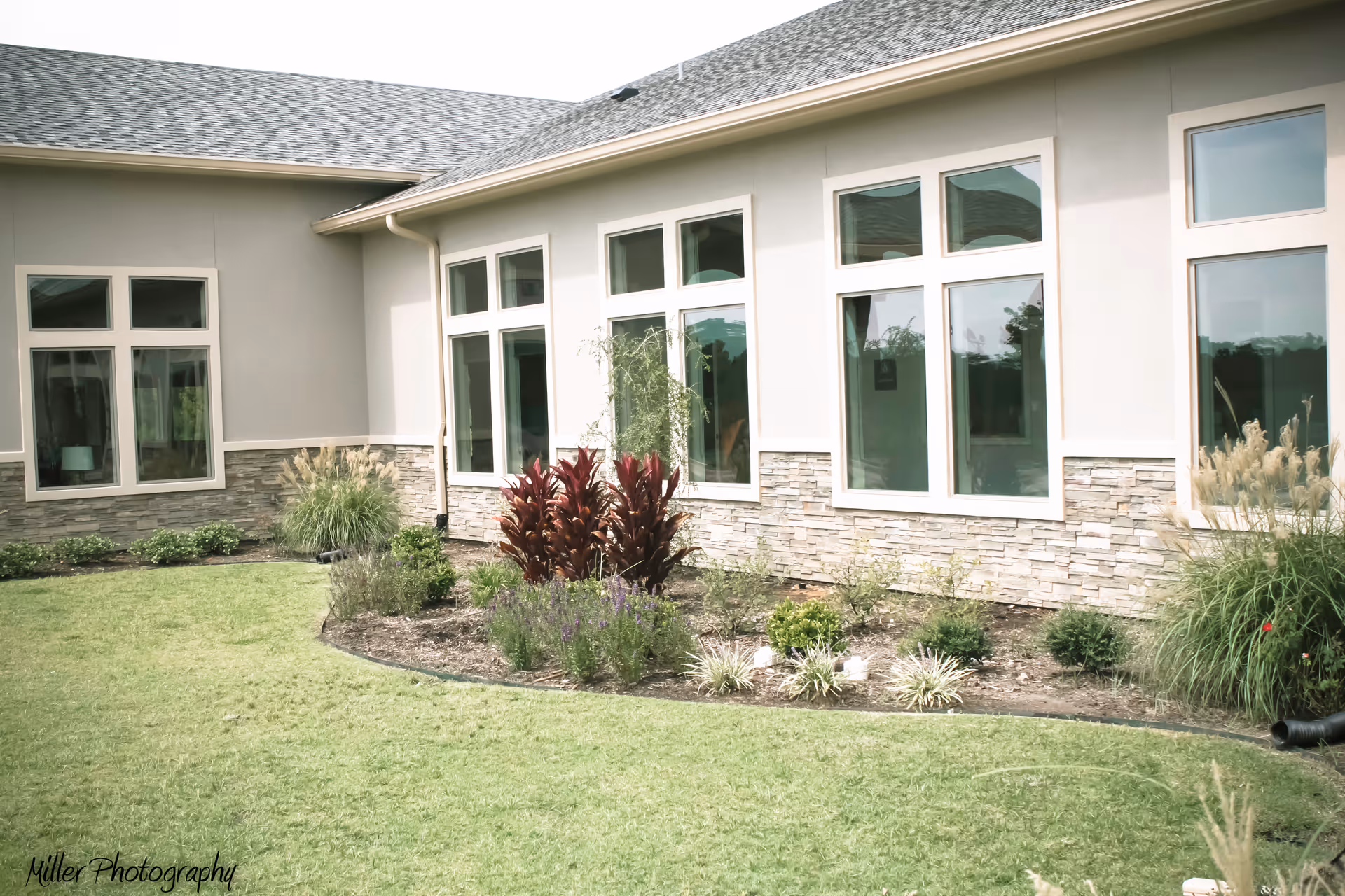 Exterior view of a single-story building with multiple large windows, beige walls with stone accents, a gray shingled roof, and a landscaped garden with green grass, shrubs, and ornamental plants in front.