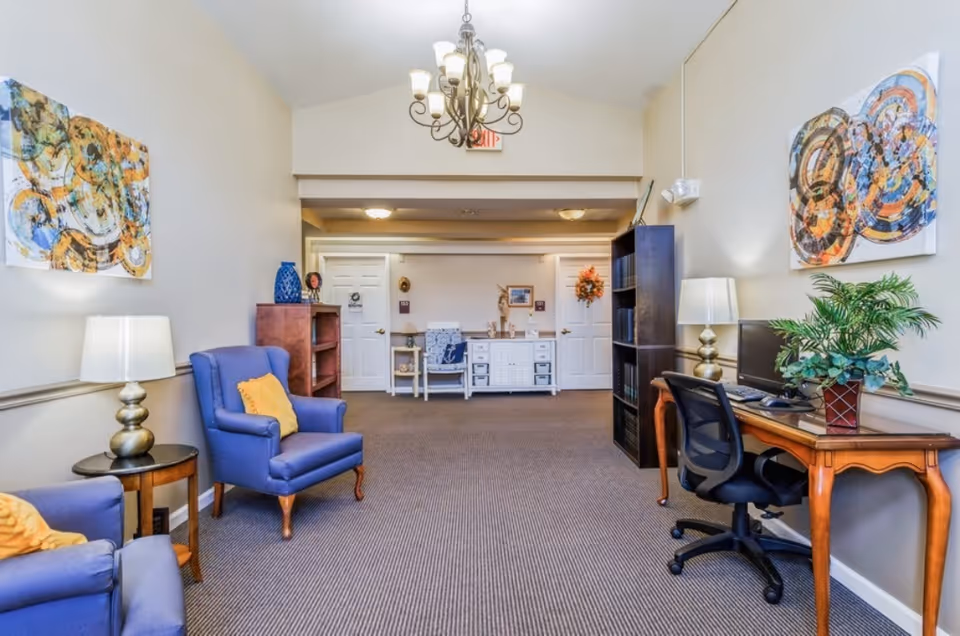 A well-lit interior room with beige walls and carpeted floor, featuring two blue armchairs with yellow pillows on the left side, a wooden side table with a lamp, and a wooden desk with a computer, office chair, lamp, and plant on the right side. There are colorful abstract paintings on the walls and a chandelier hanging from the ceiling. In the background, there are white doors and a small white cabinet with decorative items.