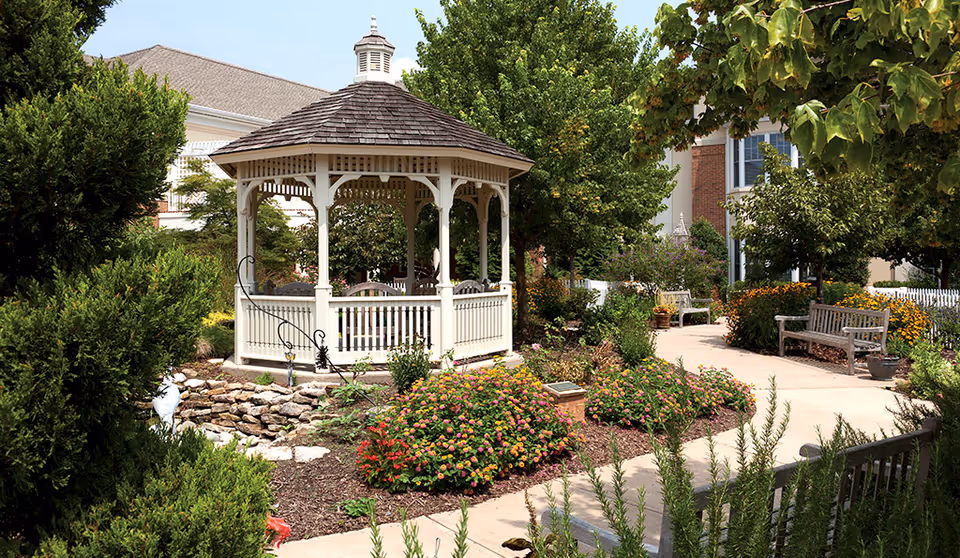 A peaceful outdoor garden area featuring a white wooden gazebo with a shingled roof, surrounded by colorful flower beds, green shrubs, and trees. There are paved walkways and wooden benches along the paths, with a building partially visible in the background.