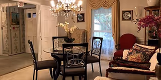 A cozy living room area with a glass-top dining table surrounded by four chairs, a chandelier overhead, a red armchair with decorative pillows, floral arrangements, and windows with curtains letting in natural light.
