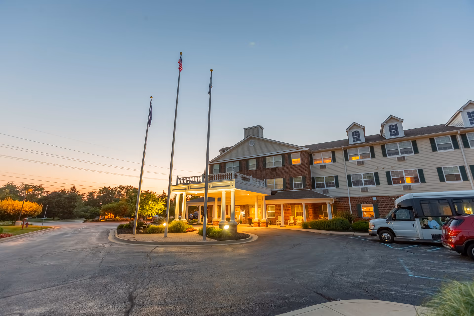 Front entrance of a multi-story senior living building at dusk with a lit porte-cochere, flagpoles, and parked vehicles.