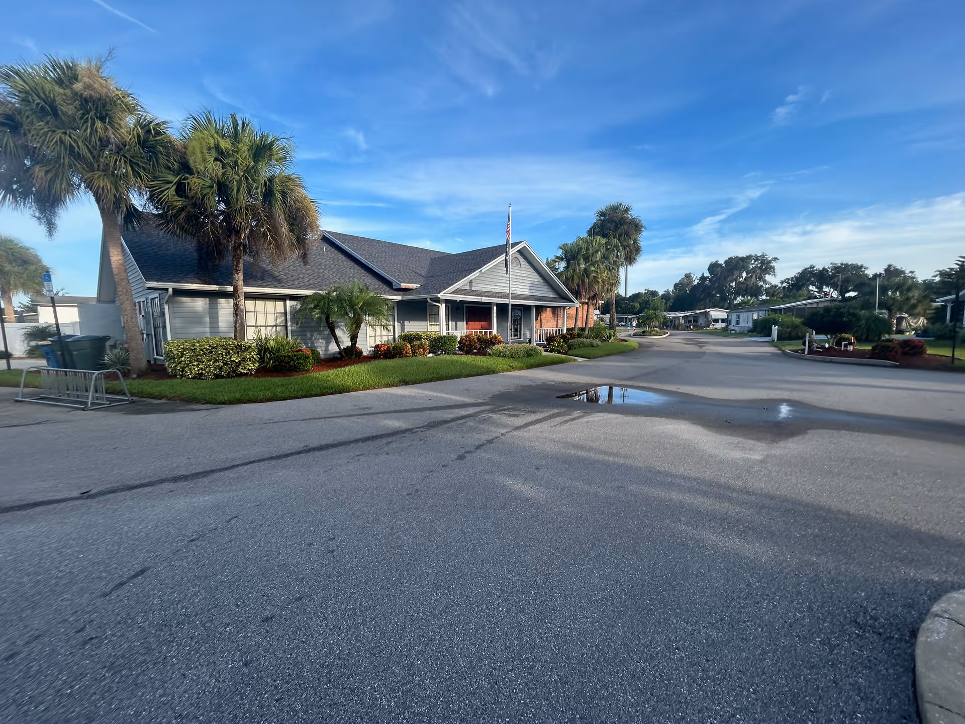 Exterior view of a single-story building with a gray roof and light gray siding, surrounded by palm trees and landscaped bushes under a blue sky with some clouds. The building is part of a retirement community, with a paved road in front and a small puddle on the road.