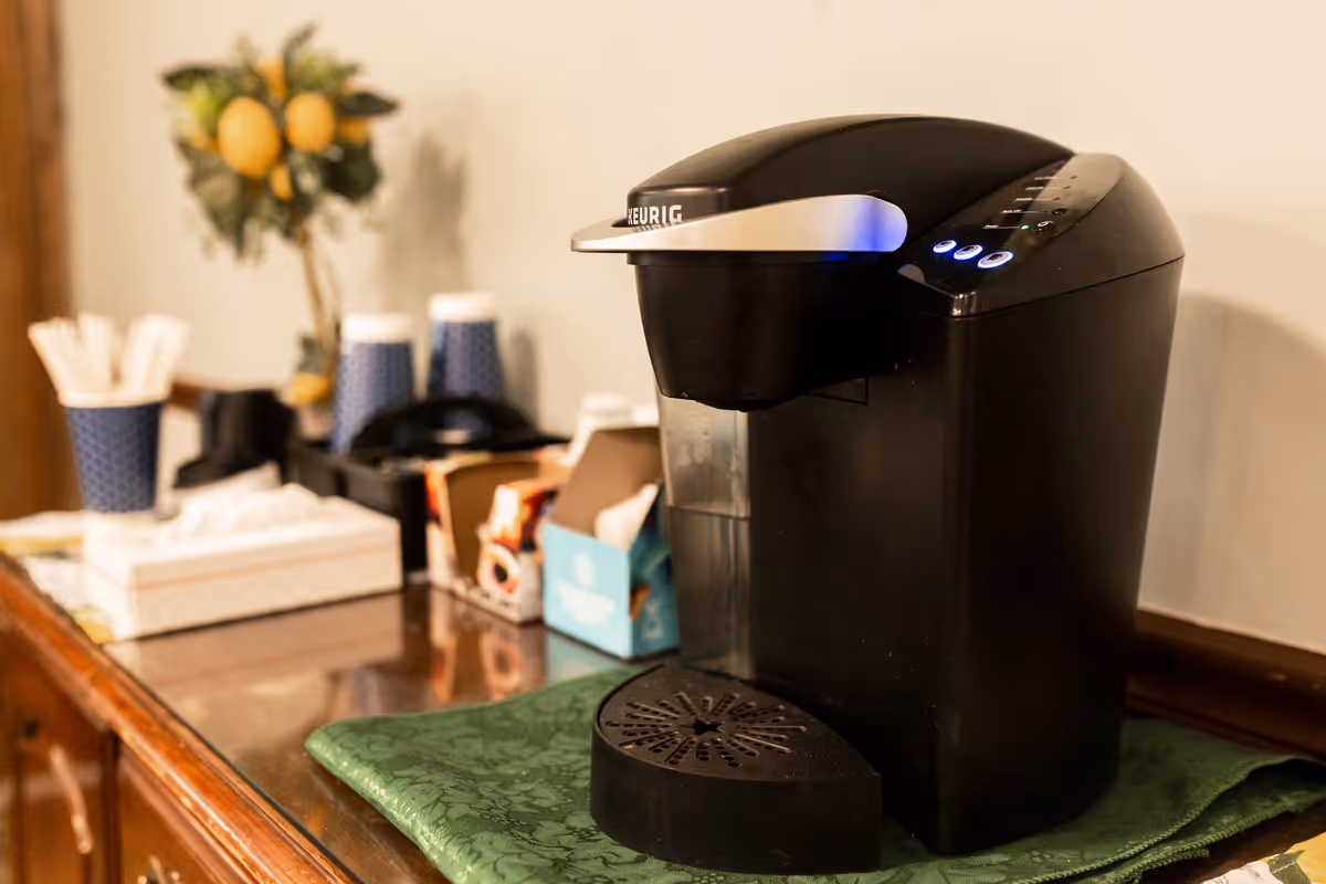 A black Keurig coffee maker on a wooden surface with a green cloth underneath. In the background, there are blue disposable cups, a box of tissues, coffee pods, and a small decorative plant with yellow fruit.