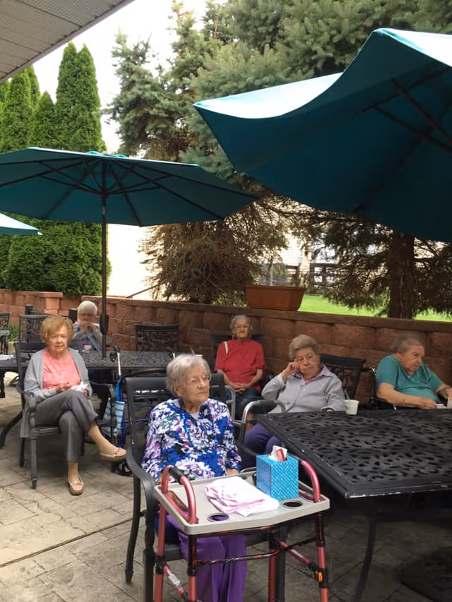A group of elderly women sitting outdoors under large teal patio umbrellas at Alexandria Manor. They are seated around black metal tables on a paved patio area with trees and a brick wall in the background. One woman in the foreground has a walker with a tray holding tissues and a cloth.