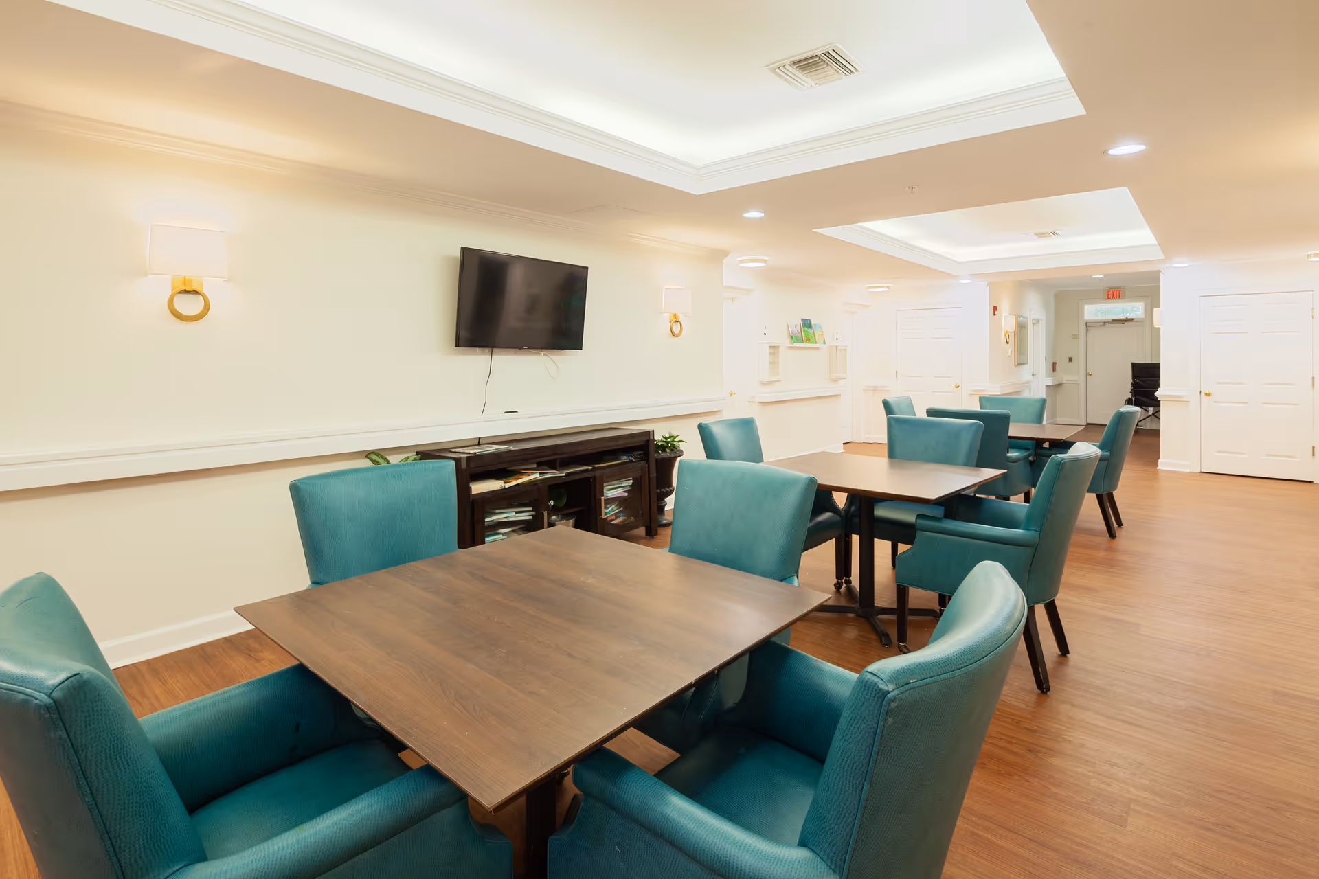 A bright and clean common area with several wooden tables surrounded by teal upholstered chairs. A flat-screen TV is mounted on the wall above a dark wooden cabinet. The room has light-colored walls, wood flooring, and recessed lighting in the ceiling.