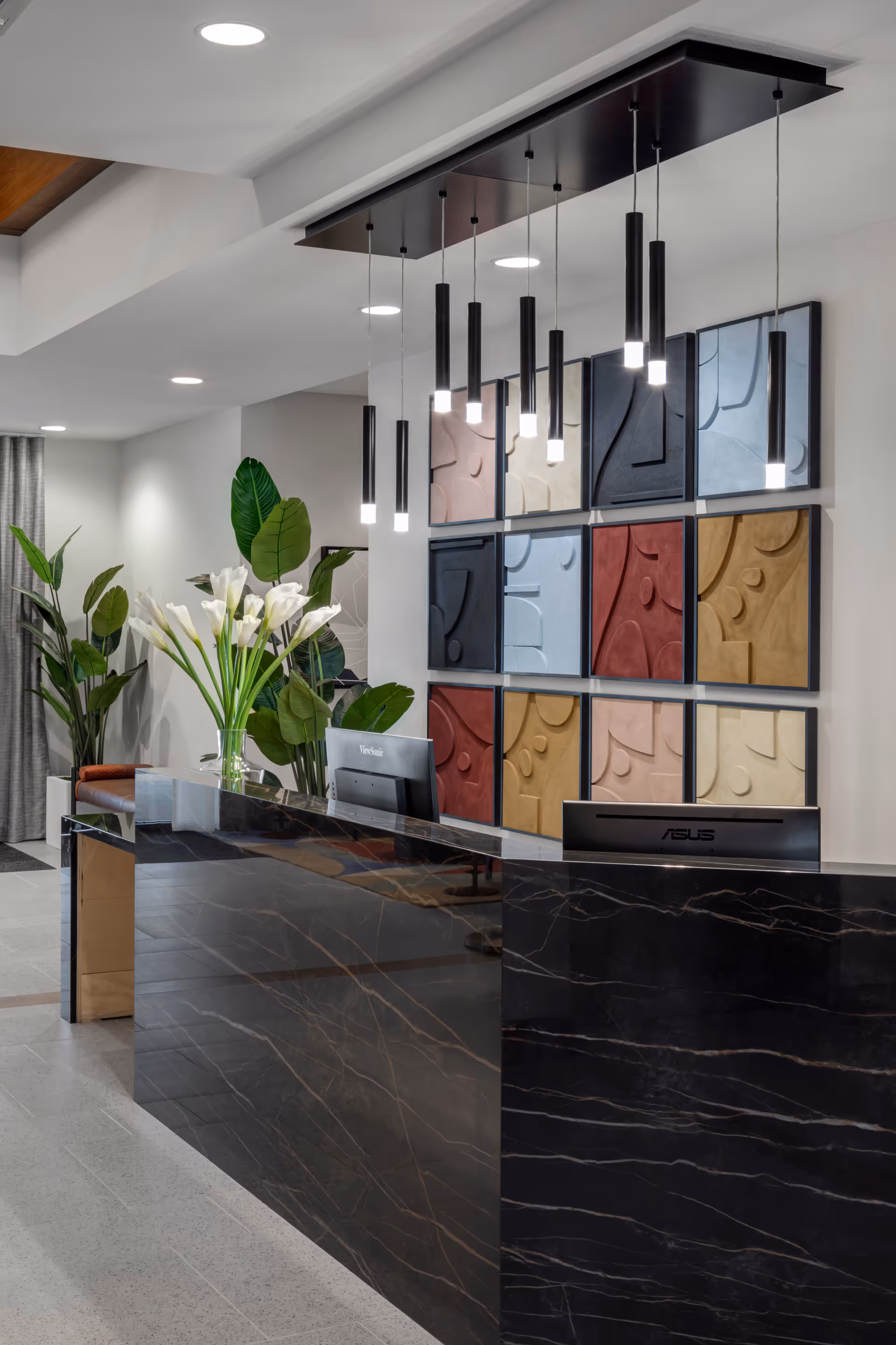 Modern reception desk area with a black marble counter, two computer monitors, and a vase of white calla lilies. Behind the desk is a wall decorated with colorful abstract square panels and hanging pendant lights. There are also large green plants in the background.