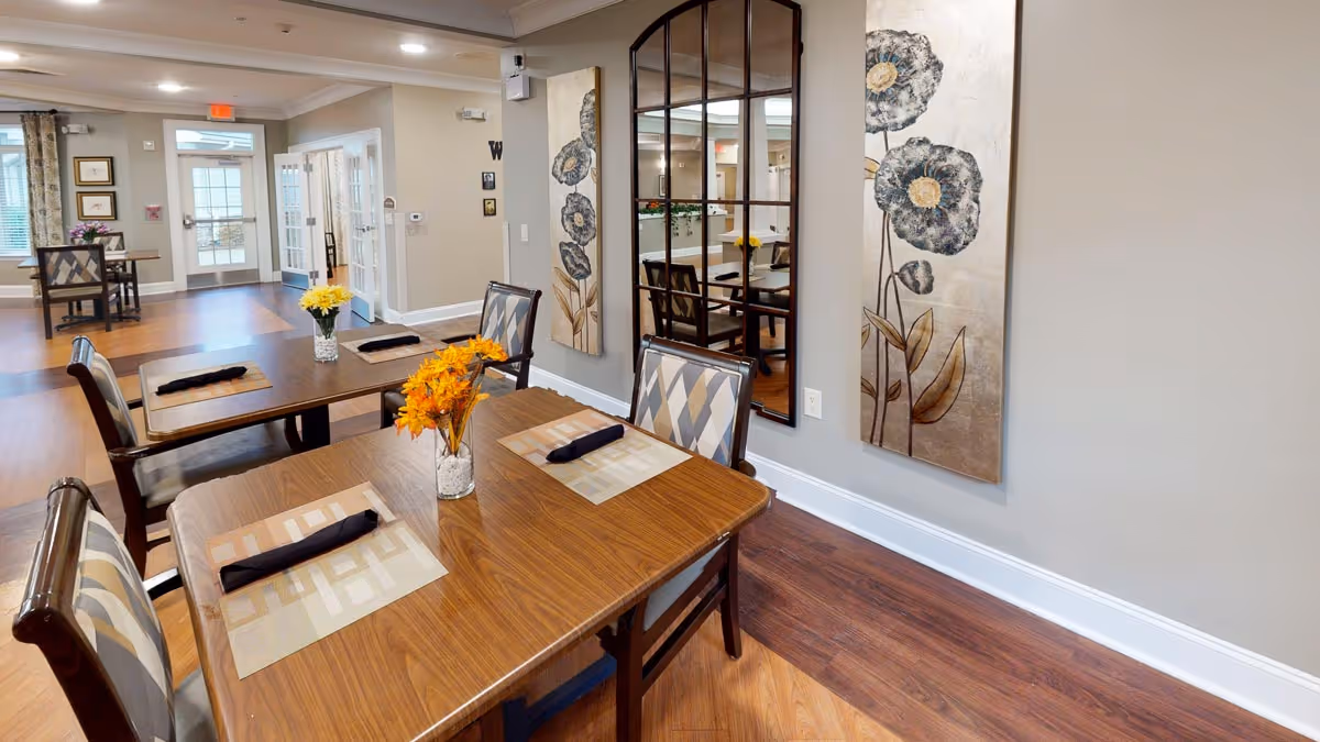 Interior dining area with wooden tables and chairs, each table set with placemats and black napkins. Vases with yellow and orange flowers are placed on the tables. The room has wooden flooring, light-colored walls, decorative floral paintings, and a large mirror on one wall. In the background, there are windows and a door leading outside.
