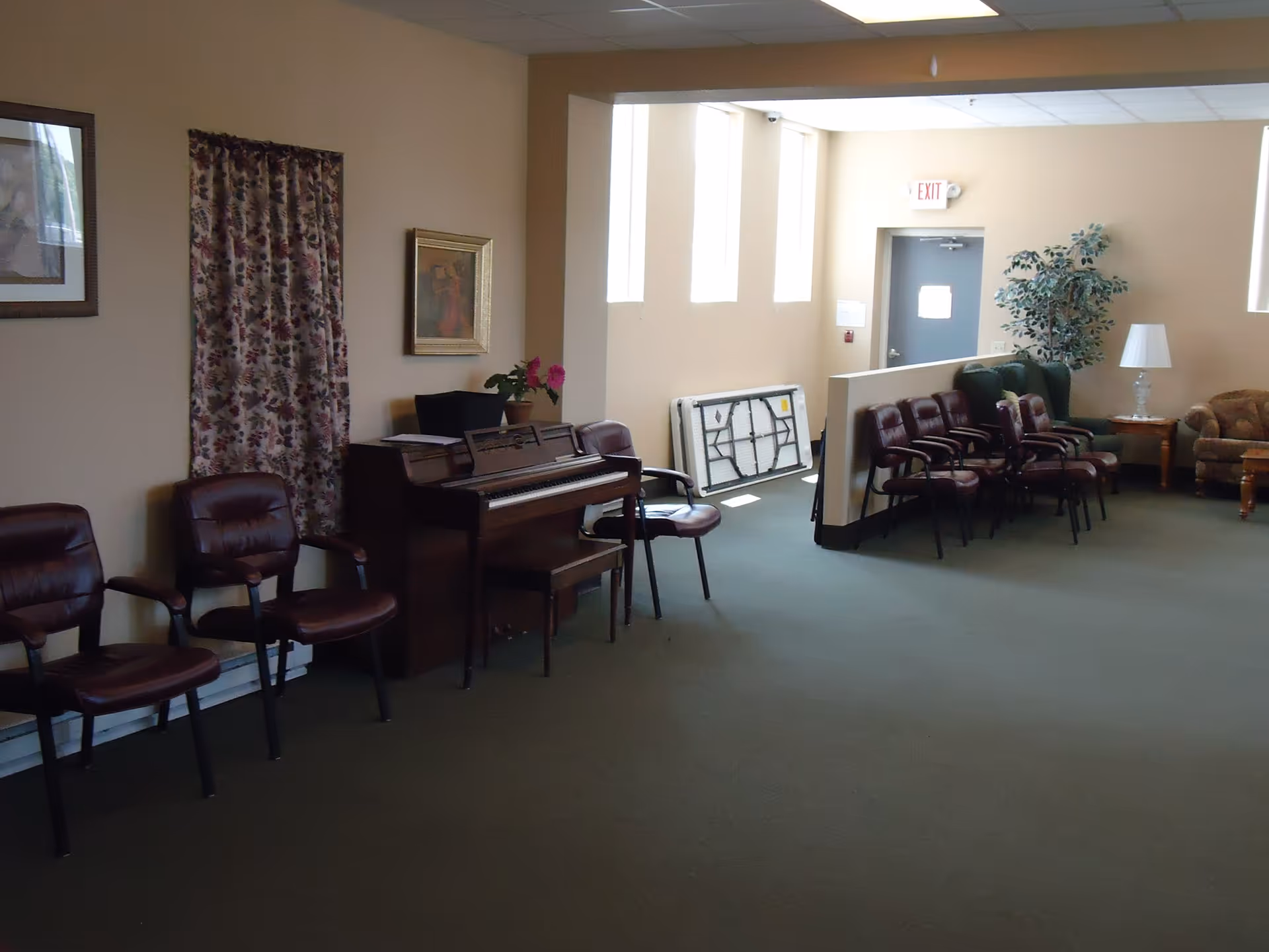 A spacious common room with an upright piano, rows of chairs, a couch, lamp and an exit door.