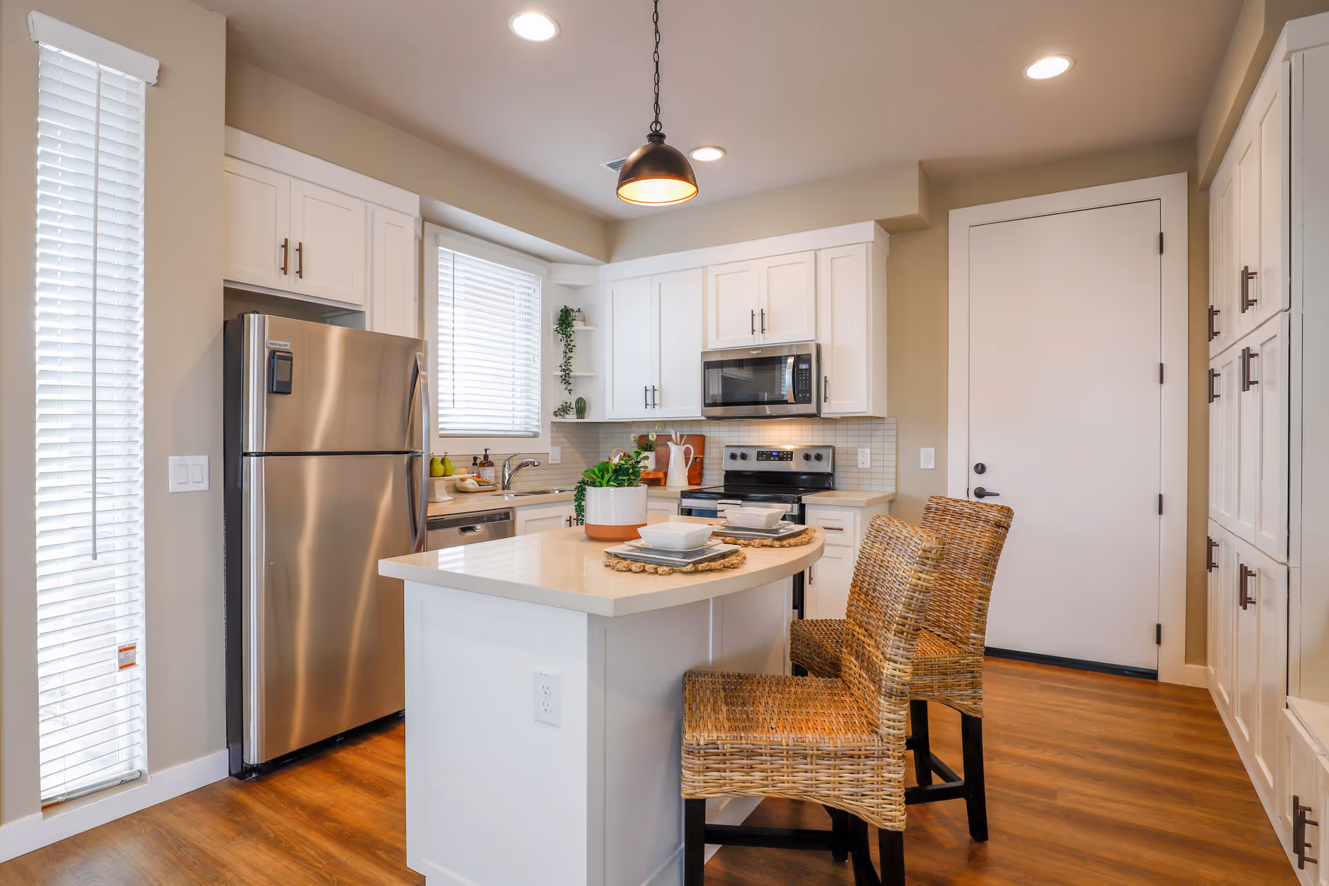 Modern kitchen with white cabinets, stainless steel refrigerator, microwave, and stove. There is a kitchen island with a light countertop, two wicker chairs, and place settings. The floor is wooden, and there is a window with blinds letting in natural light.