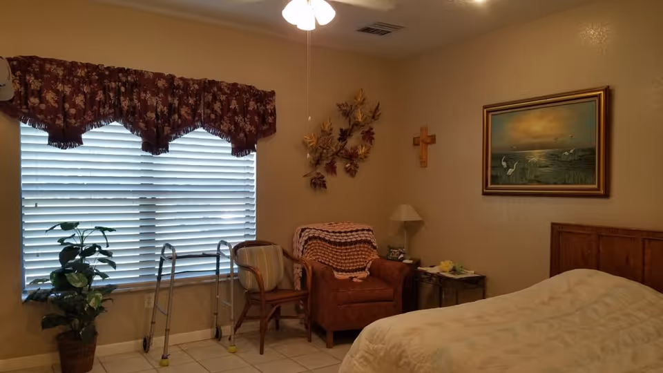 Assisted living bedroom with a bed, armchair draped with a blanket, walker, potted plant, and a window with blinds.