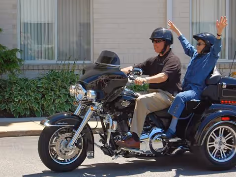 Two people riding a black three-wheeled motorcycle in front of a building with large windows and green plants. The driver is wearing a black helmet and sunglasses, while the passenger, also wearing a helmet and sunglasses, has their arms raised in excitement.