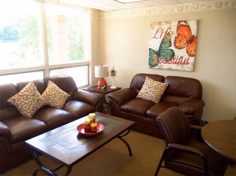A cozy living room with two brown leather sofas adorned with patterned throw pillows, a wooden coffee table with a red tray holding three candles, a side table with a lamp and a small decorative birdhouse, and a wall art featuring colorful butterflies and the words 'Life Beautiful'. Large windows allow natural light to fill the room.