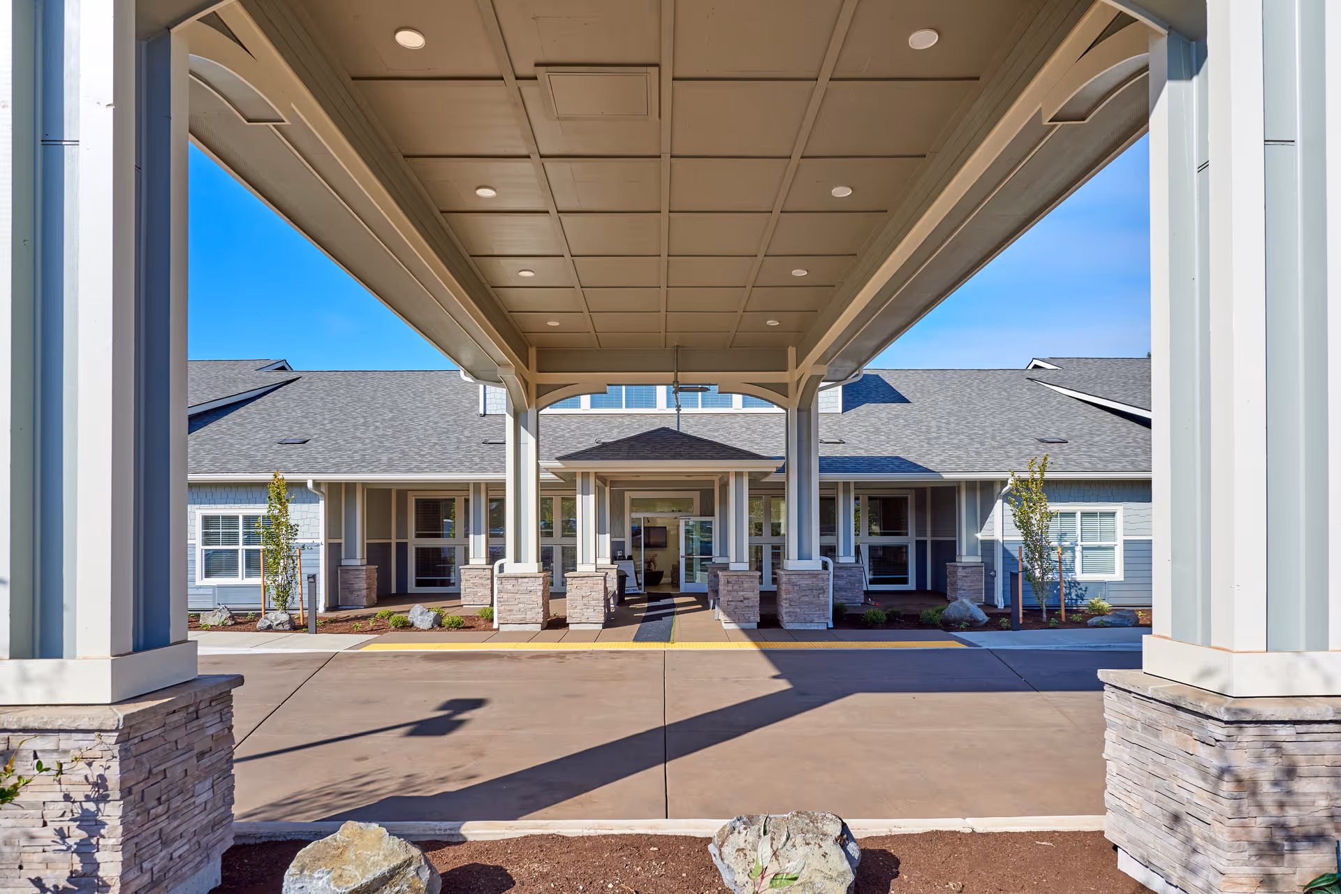 View of the covered entrance area of a senior living facility with stone pillars and a paved driveway leading to the front door under a clear blue sky.