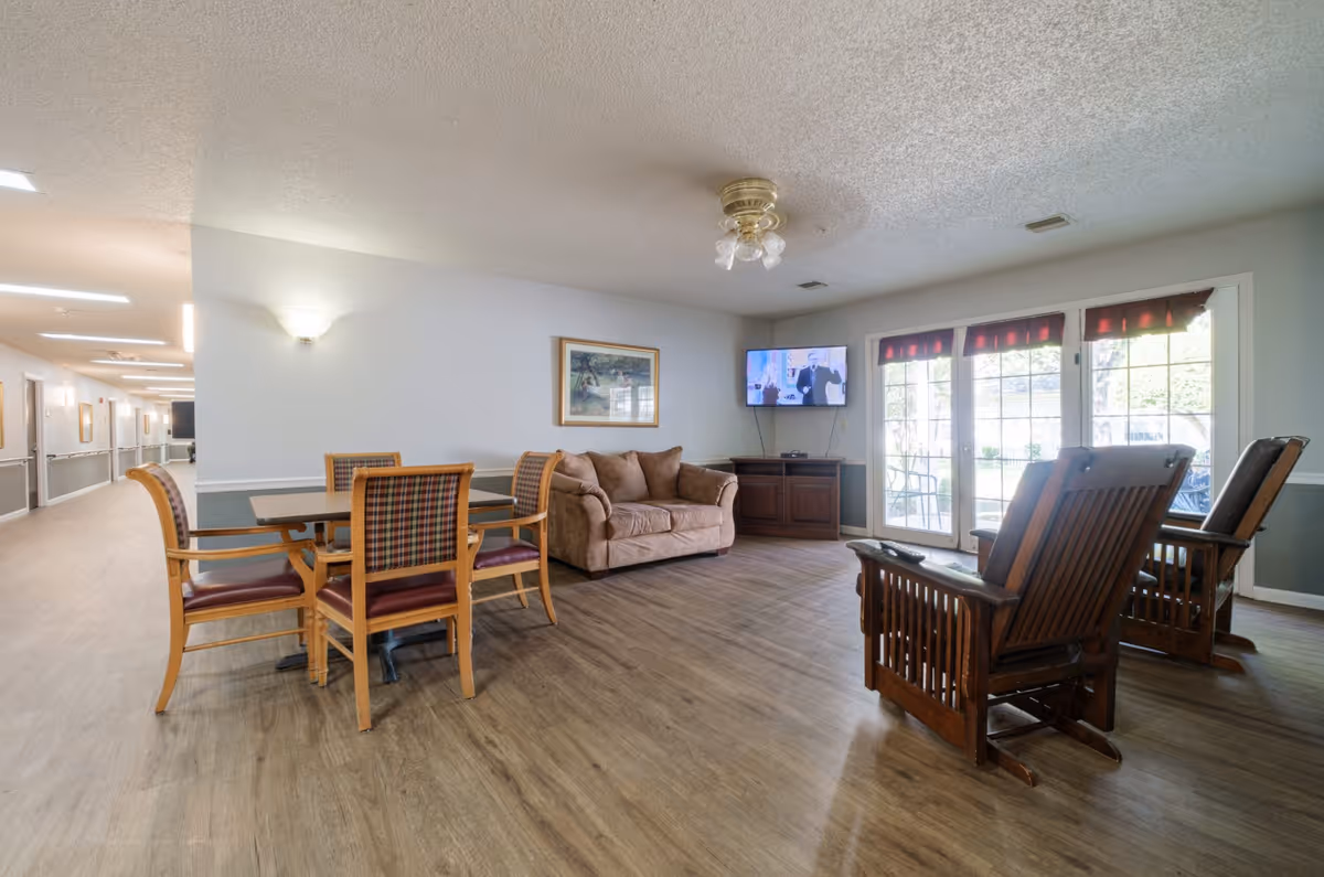 Spacious senior living common room with a dining table and chairs, a sofa, wooden armchairs, a wall-mounted TV, and large windows/doors.