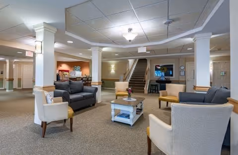 Spacious senior living lounge with multiple seating areas, a central coffee table, columns and a staircase in the background.