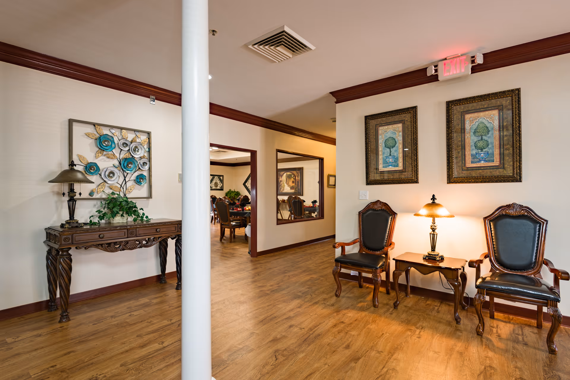 Interior view of a hallway in Brentwood Retirement Community featuring wooden flooring, a decorative wooden console table with a lamp and plant, two ornate chairs with a small table and lamp between them, framed artwork on the walls, and an exit sign above a doorway leading to a dining area with tables and chairs.