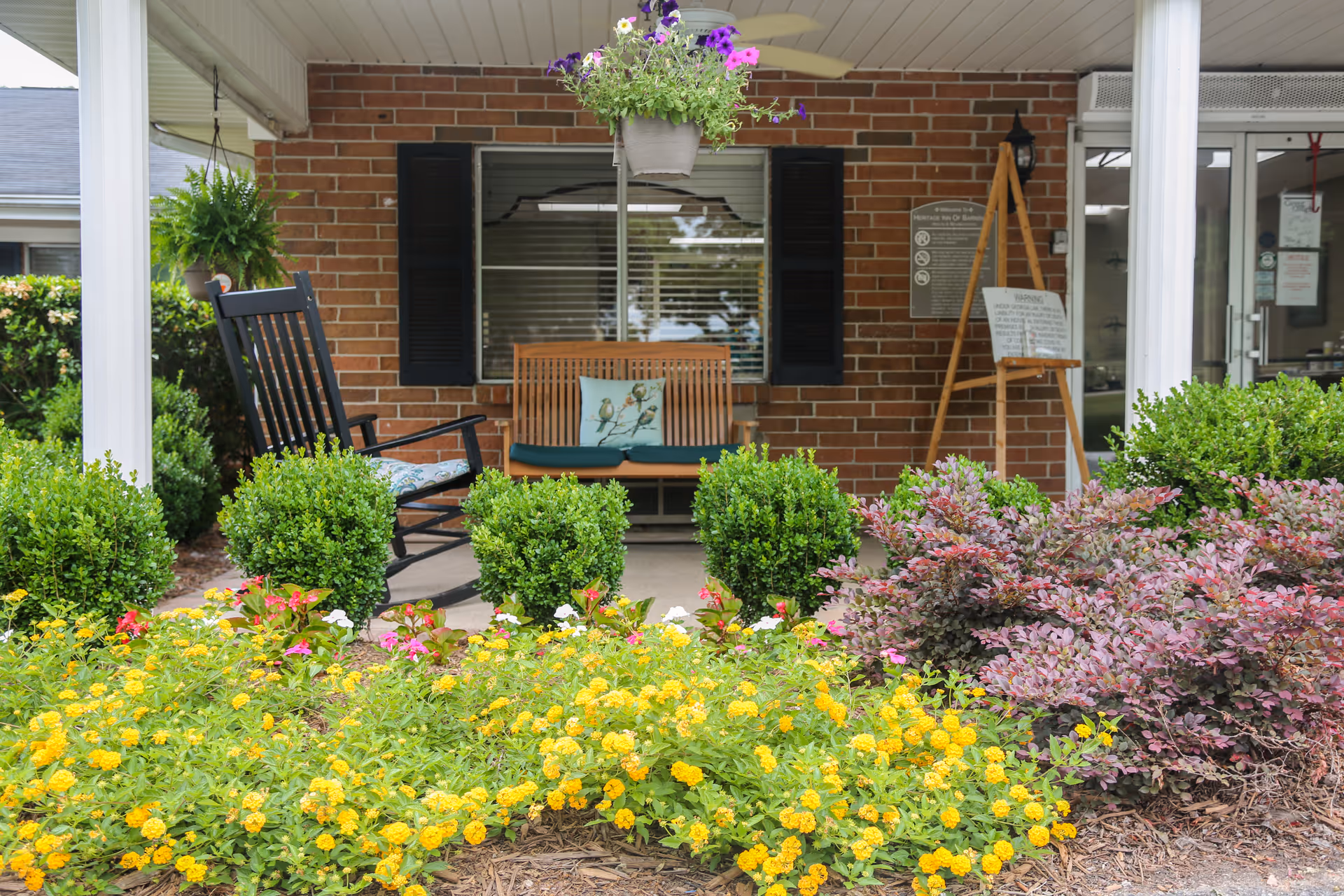 Front porch area of a building with a brick wall, featuring a wooden bench with a decorative pillow, a black rocking chair with a cushion, hanging flower pots, and well-maintained garden beds with yellow and purple flowers and green shrubs.