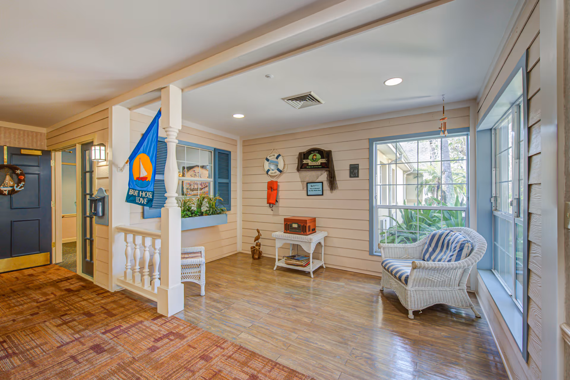 A cozy indoor seating area with nautical-themed decor including a blue flag with a sailboat, a life preserver, and a wooden pelican sculpture. The space features a wicker loveseat with striped cushions, a small white wicker table with a vintage-style radio, large windows letting in natural light, and a blue door leading to another room.