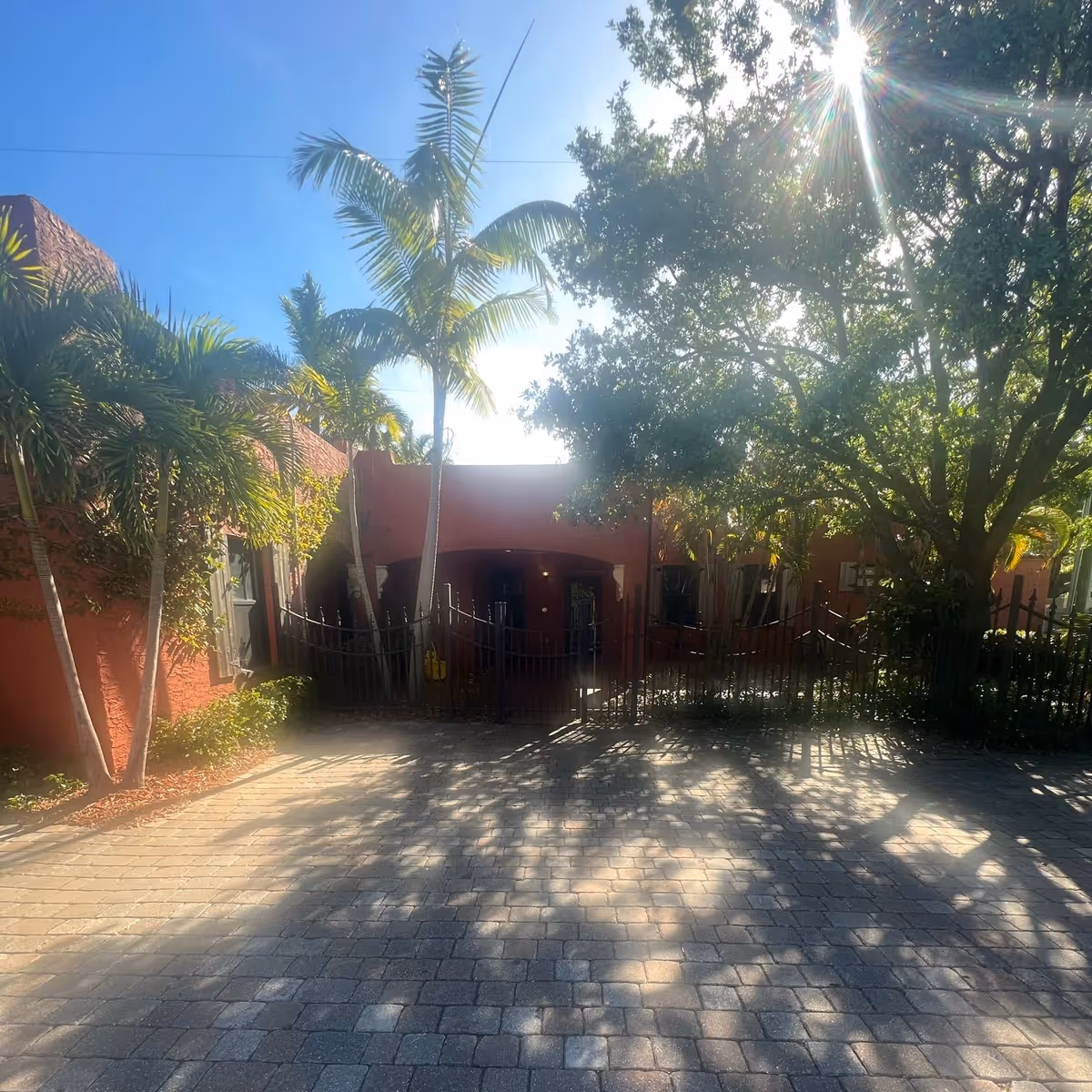 Sunlit front entrance of a red stucco building with palm trees, a black gate, and patterned driveway shadows.