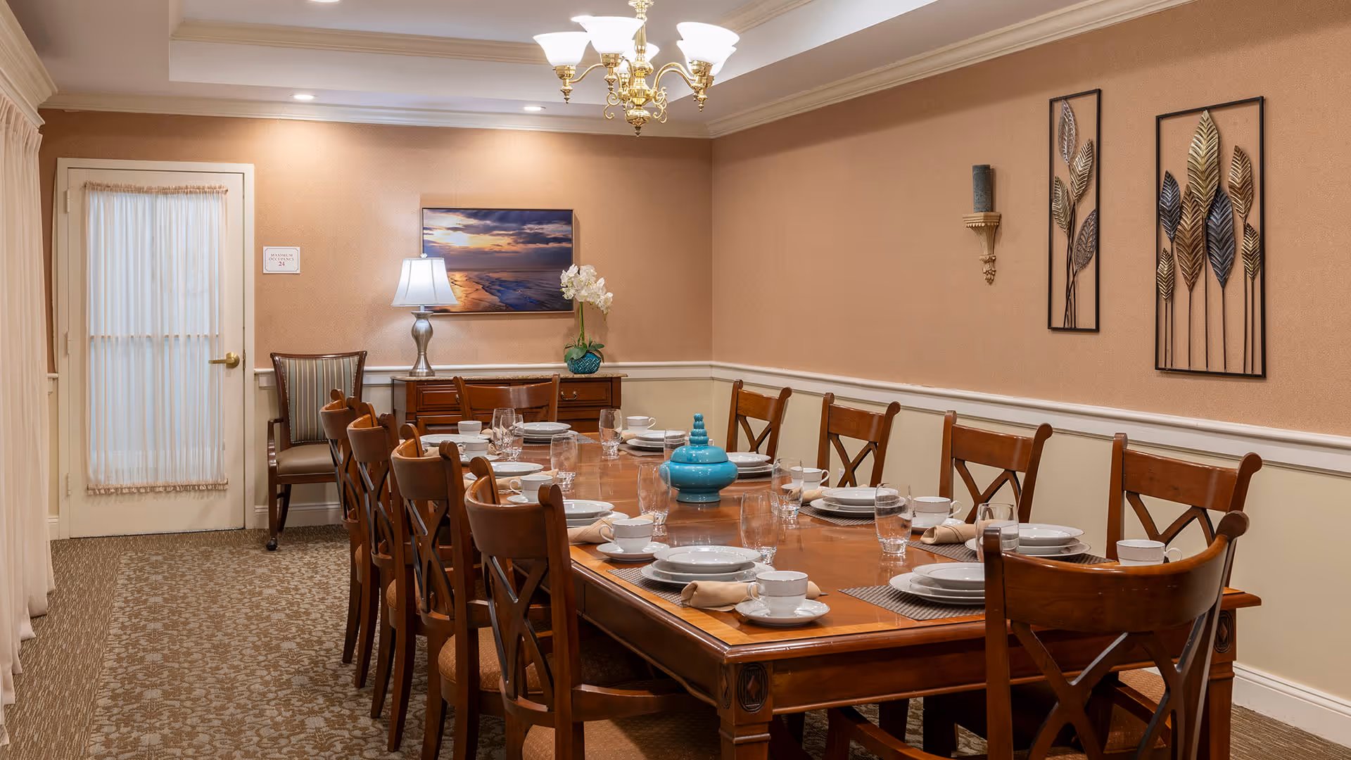 A formal dining room with a long wooden table set for twelve people. The table is arranged with white plates, bowls, cups, glasses, and beige napkins on placemats. The room has beige walls with white wainscoting, a chandelier overhead, and decorative metal leaf wall art. There is a sideboard with a lamp, a vase with white flowers, and a framed picture of a sunset over the ocean. A door with a sheer curtain is visible on the left side.