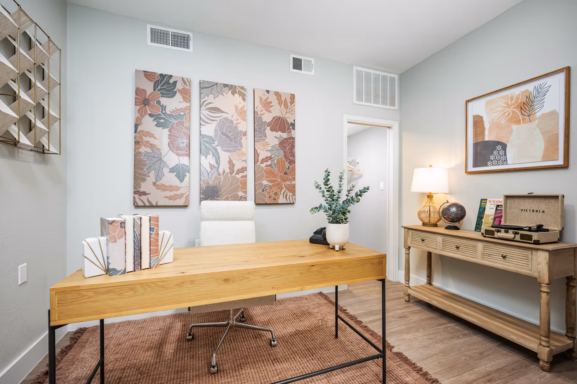 A bright furnished office with a wooden desk, floral wall art, and a console table holding a lamp and record player.