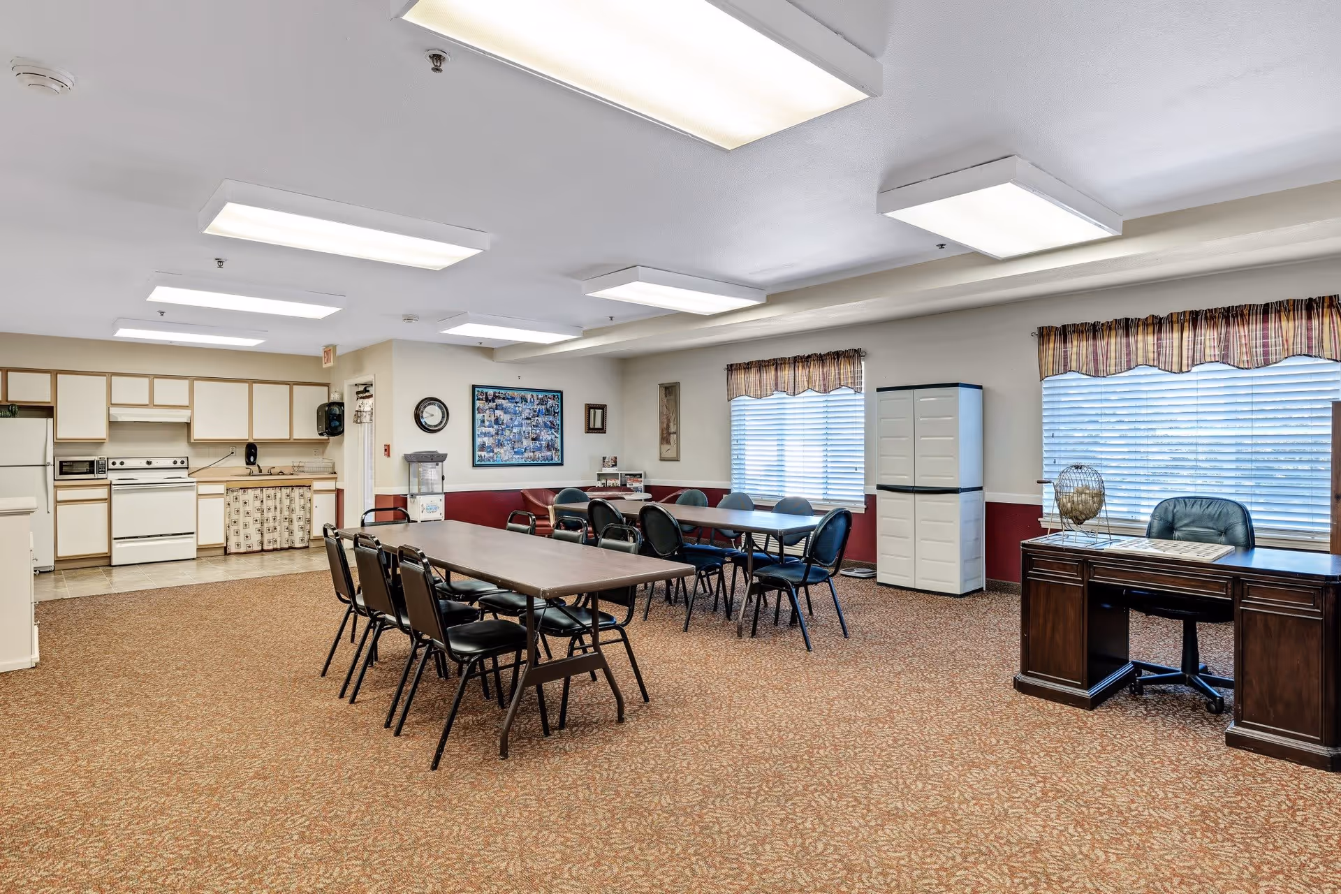 A spacious room with a kitchen area on the left featuring white cabinets, a stove, microwave, and refrigerator. In the center, there are two rectangular tables with black chairs arranged around them. On the right side, there is a dark wooden desk with a black office chair and a globe on top. The room has carpeted flooring, large windows with blinds and plaid valances, and ceiling lights.