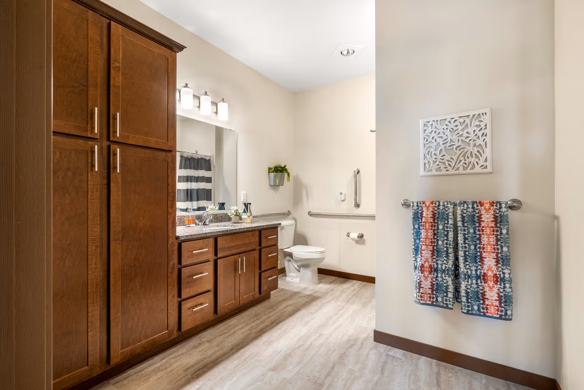 Spacious bathroom with dark wood cabinets and granite countertop, toilet with grab bars, and a colorful towel hanging on a wall rack.