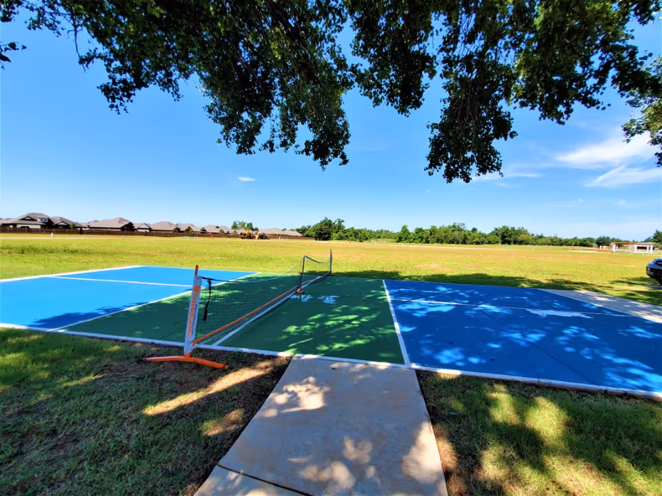 Shaded outdoor pickleball/tennis court with blue and green painted surfaces, a net, a sidewalk leading to it, and an open grassy field with houses in the distance.