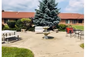 Outdoor patio area with a concrete surface featuring a central stone fountain, surrounded by benches and metal chairs with tables. In the background, there is a single-story brick building with a red roof and some greenery including a large evergreen tree and bushes.