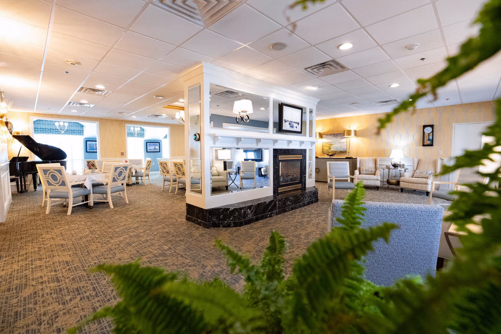 A spacious and well-lit senior living facility common area featuring a central fireplace with mirrors, several seating arrangements including armchairs and dining tables with white chairs, a grand piano in the corner, and decorative plants in the foreground.