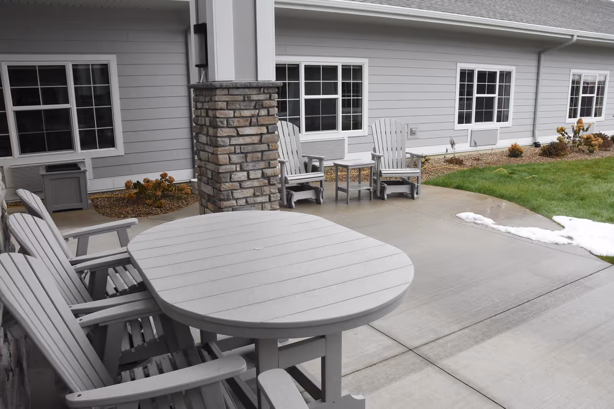 A covered outdoor patio with a round table and Adirondack-style chairs by a stone column outside a light-gray building, with grass and a small patch of snow.