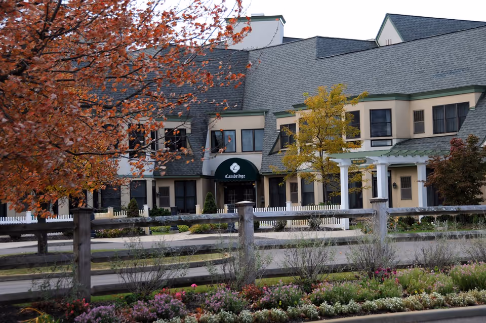Exterior view of a senior living facility building with beige walls and green roof trim. There is a green awning with the word 'Cambridge' above the entrance. The foreground shows a wooden fence, colorful flower beds, and trees with autumn foliage.