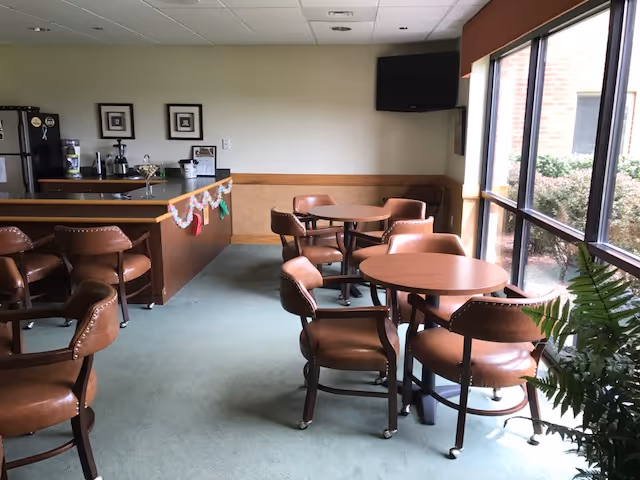 A small dining area with round wooden tables and brown leather chairs. There is a counter with bar stools on the left side, decorated with a garland. A flat-screen TV is mounted on the wall in the corner. Large windows on the right side let in natural light and show some greenery outside.