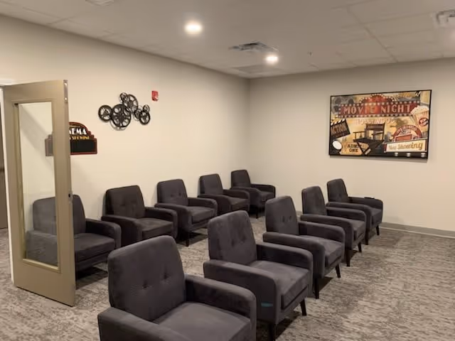 A small theater room with two rows of gray upholstered armchairs arranged facing a blank wall. The room has a gray carpet, white walls, and a ceiling with recessed lighting. On the wall to the right, there is a framed vintage-style movie poster. On the left wall near an open door, there is a decorative metal wall art piece and a small sign that reads 'Cinema Screening'.