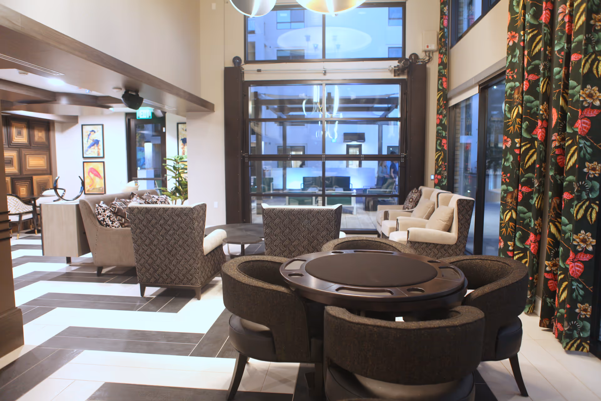 A modern and stylish living room area in Clarendale Arcadia featuring a round table with four cushioned chairs in the foreground, multiple armchairs and sofas arranged around a low coffee table, large windows with floral curtains, and decorative artwork on the walls.