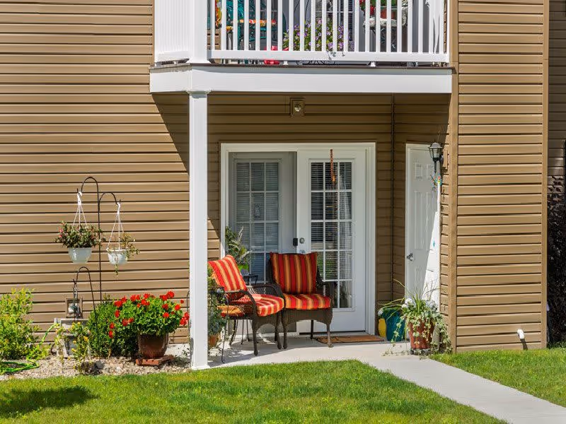 Outdoor patio area of a residential building with two striped cushioned chairs, potted plants, hanging flower pots, and a white railing balcony above. The building has beige siding and white framed glass doors leading inside.