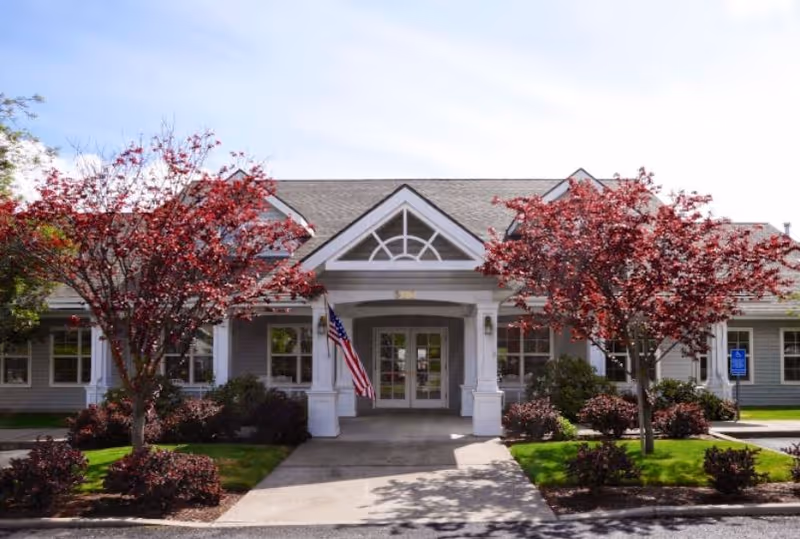Front entrance of a single-story senior living building with a covered portico, an American flag, and landscaped red-leaf trees.