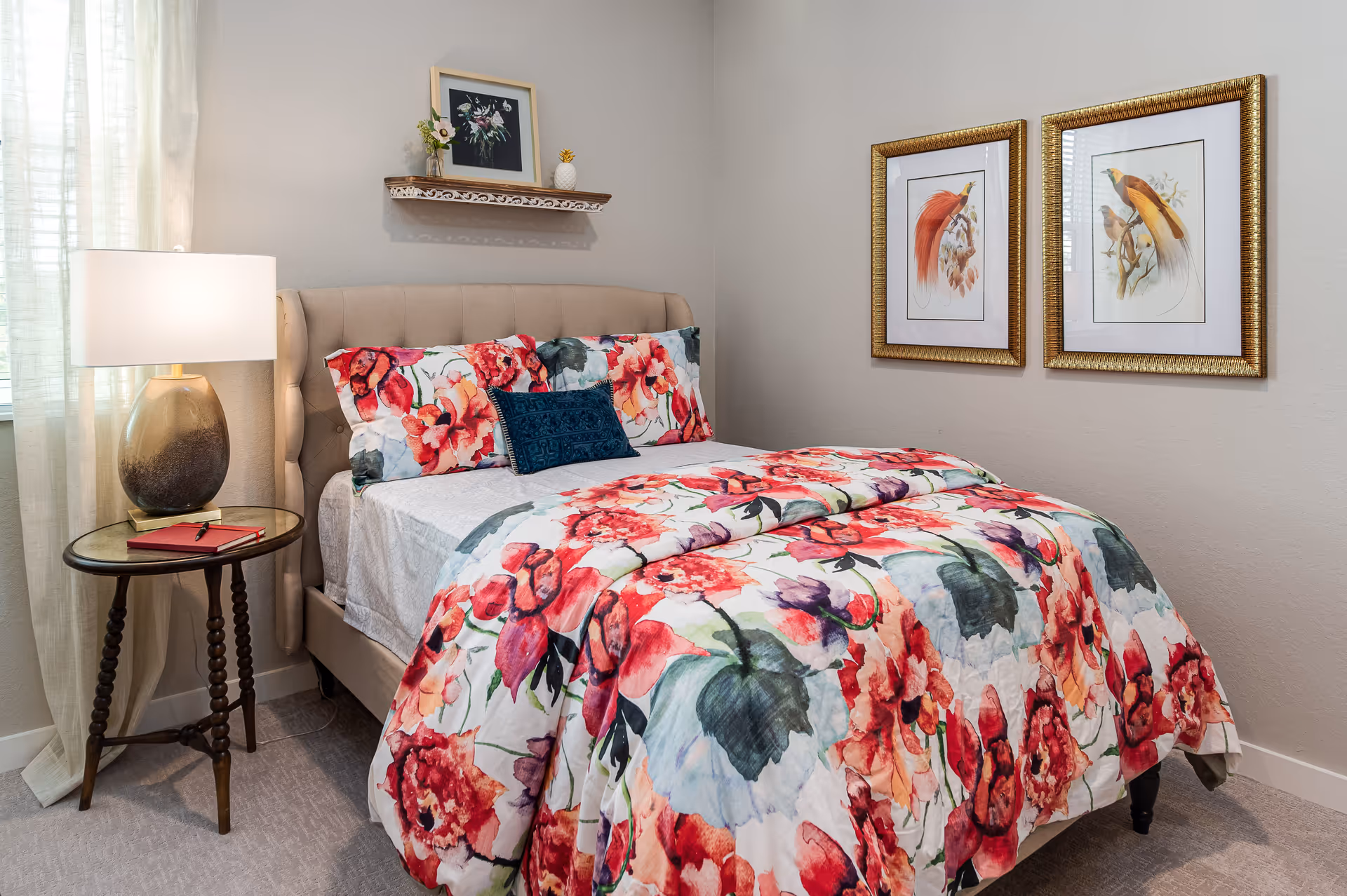 A cozy bedroom with a beige upholstered bed featuring a colorful floral bedspread and matching pillows. A small round wooden side table with a lamp and a red notebook is placed next to the bed. On the wall above the bed is a small shelf with a framed floral picture and decorative items. Two framed bird illustrations hang on the adjacent wall.