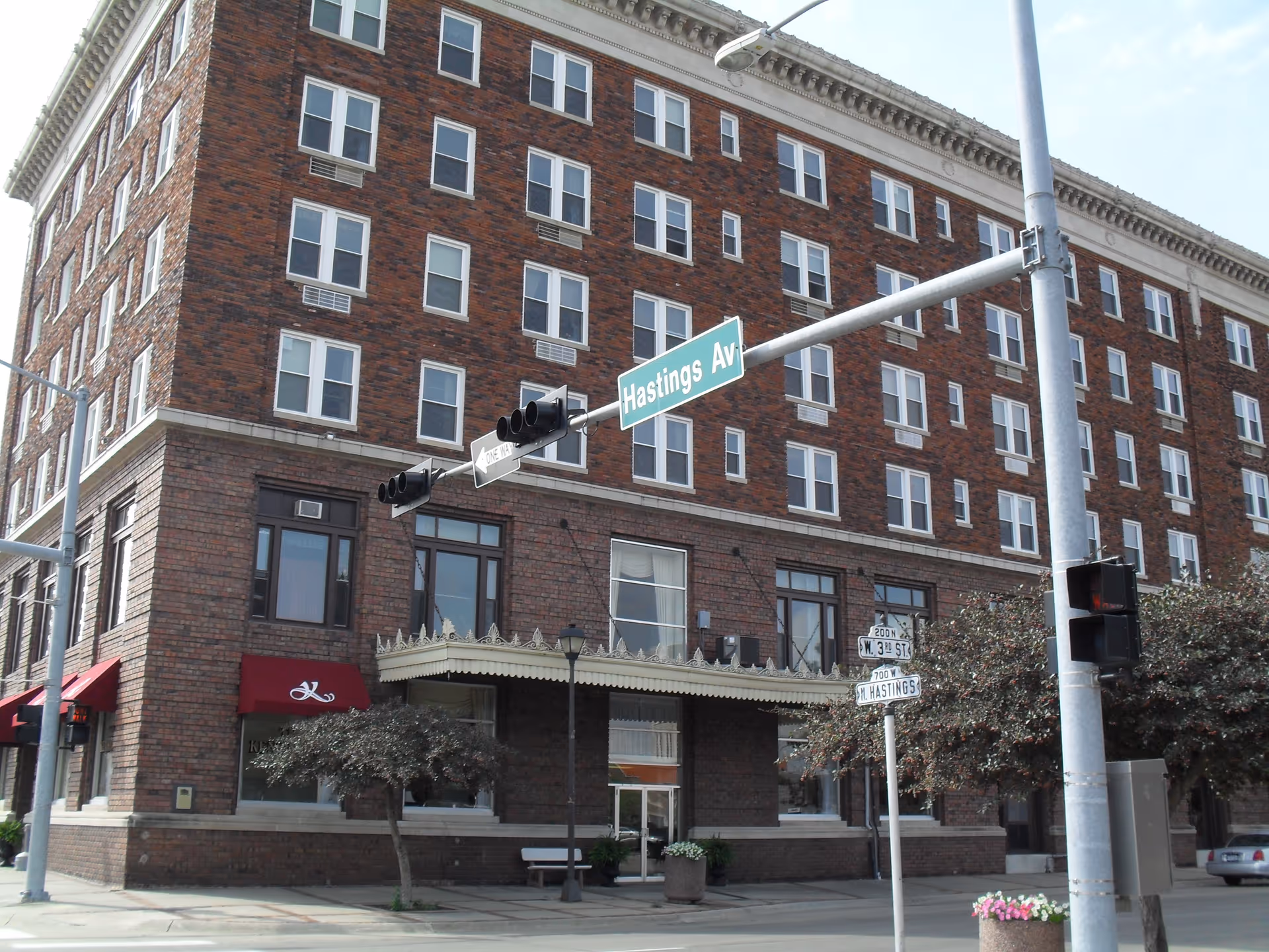 Exterior view of a multi-story brick building on a street corner with a red awning and a sign that reads 'The Kensington'. There are traffic lights and street signs for Hastings Avenue and W. 3rd Street in front of the building.