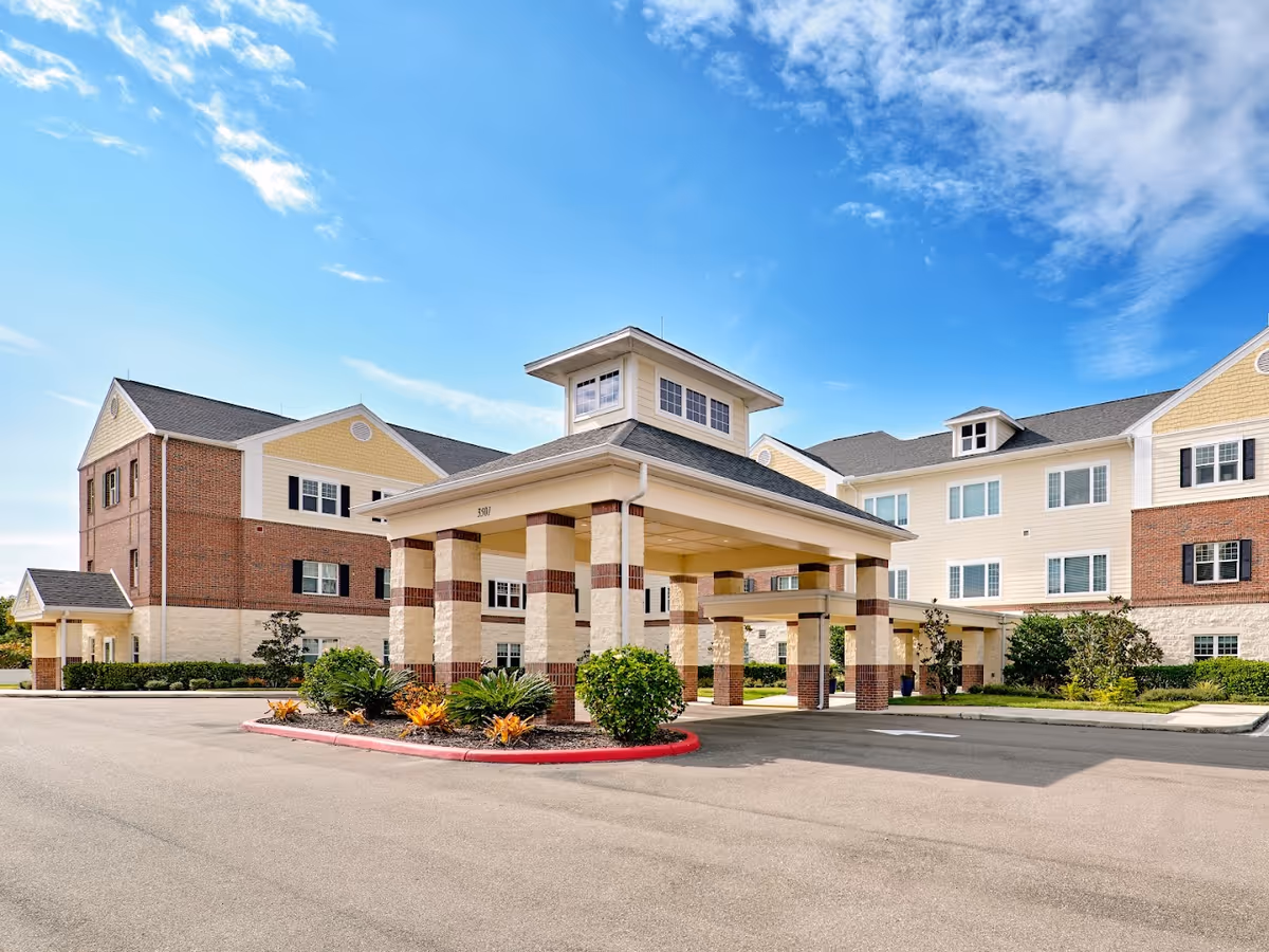 Exterior view of Volante of Fort Myers senior living facility showing a covered entrance with columns, landscaped plants, and a multi-story building with brick and light-colored siding under a blue sky with some clouds.