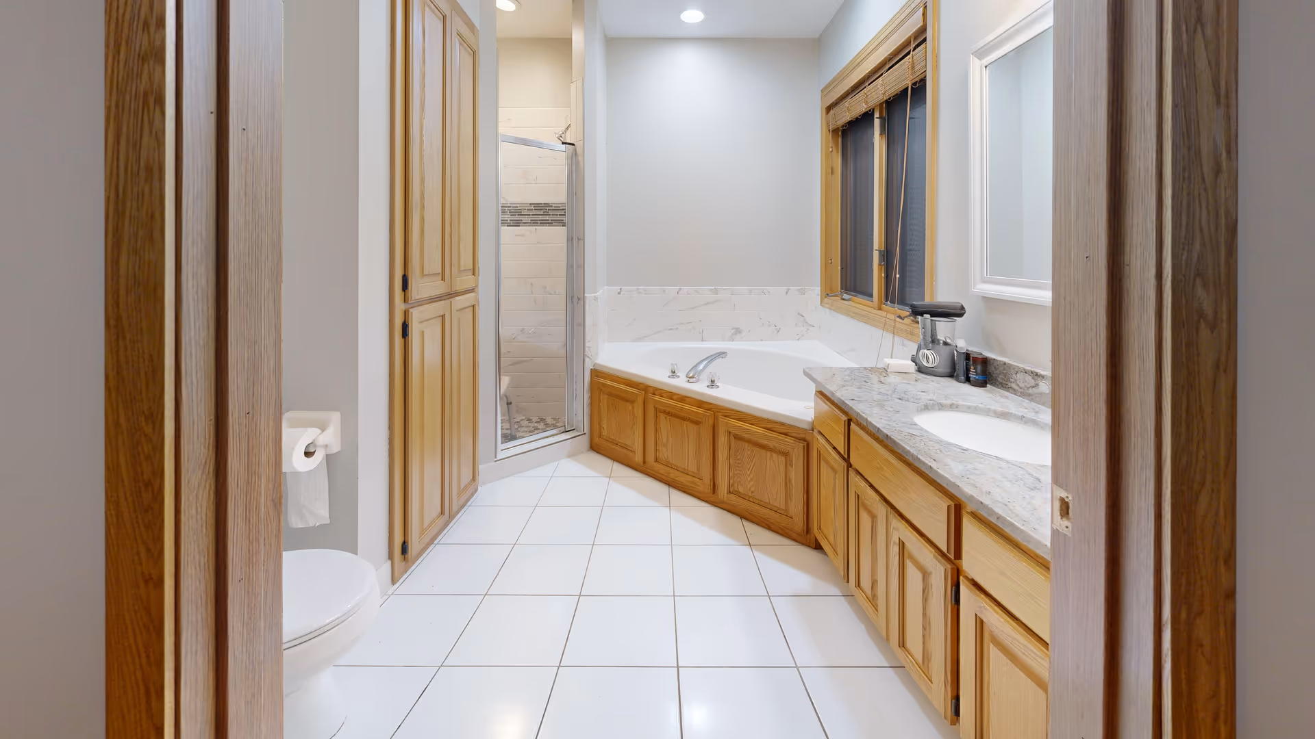 A spacious bathroom featuring a white tiled floor, a corner bathtub with wooden paneling, a glass-enclosed shower, a toilet with a roll of toilet paper, wooden cabinetry, a granite countertop with a sink, a window with wooden blinds, and a wall-mounted mirror.