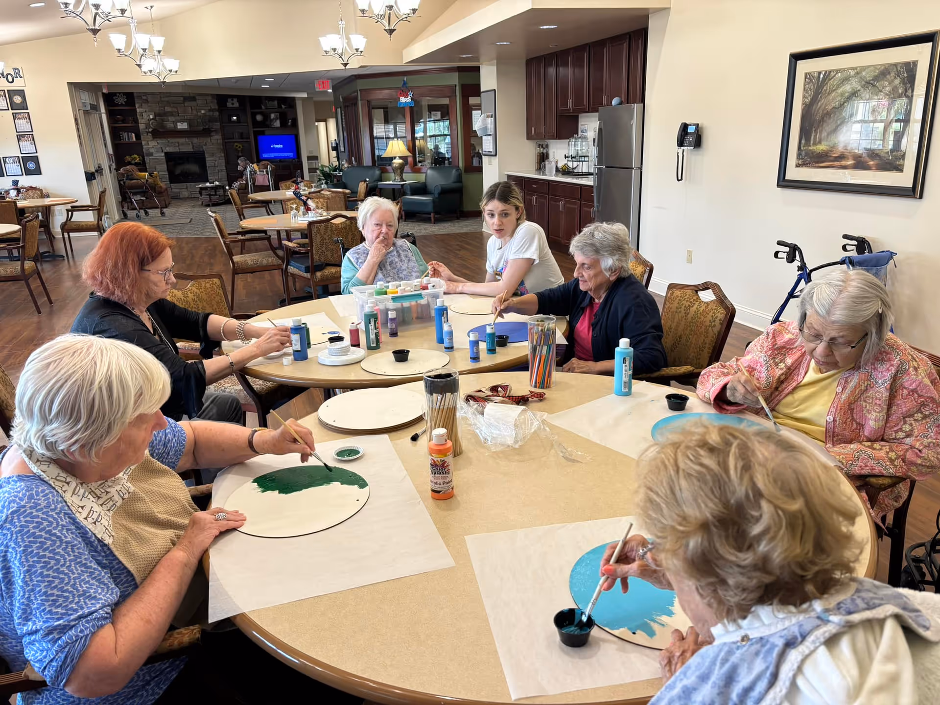 A group of elderly residents and a caregiver sit around a large communal dining/activity table painting round wooden plaques.