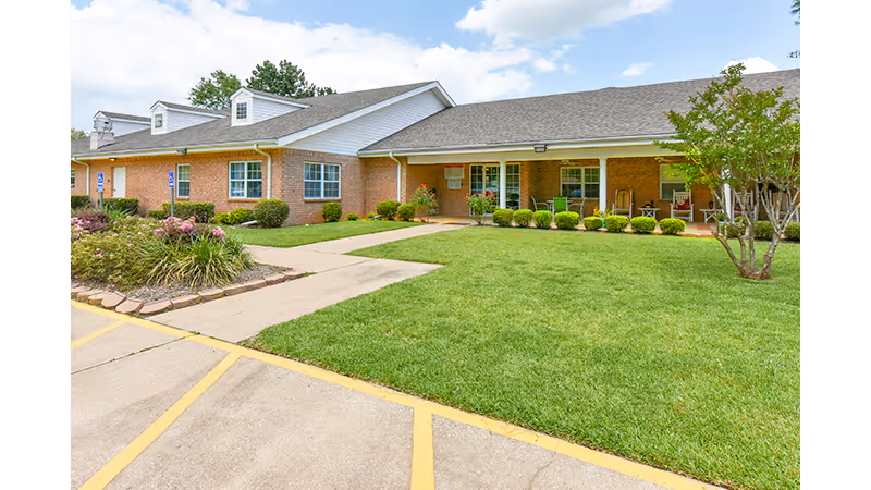 Exterior view of Azalea Place, a single-story brick building with a well-maintained lawn, shrubs, and a concrete walkway leading to the entrance. The sky is partly cloudy and the area looks clean and inviting.