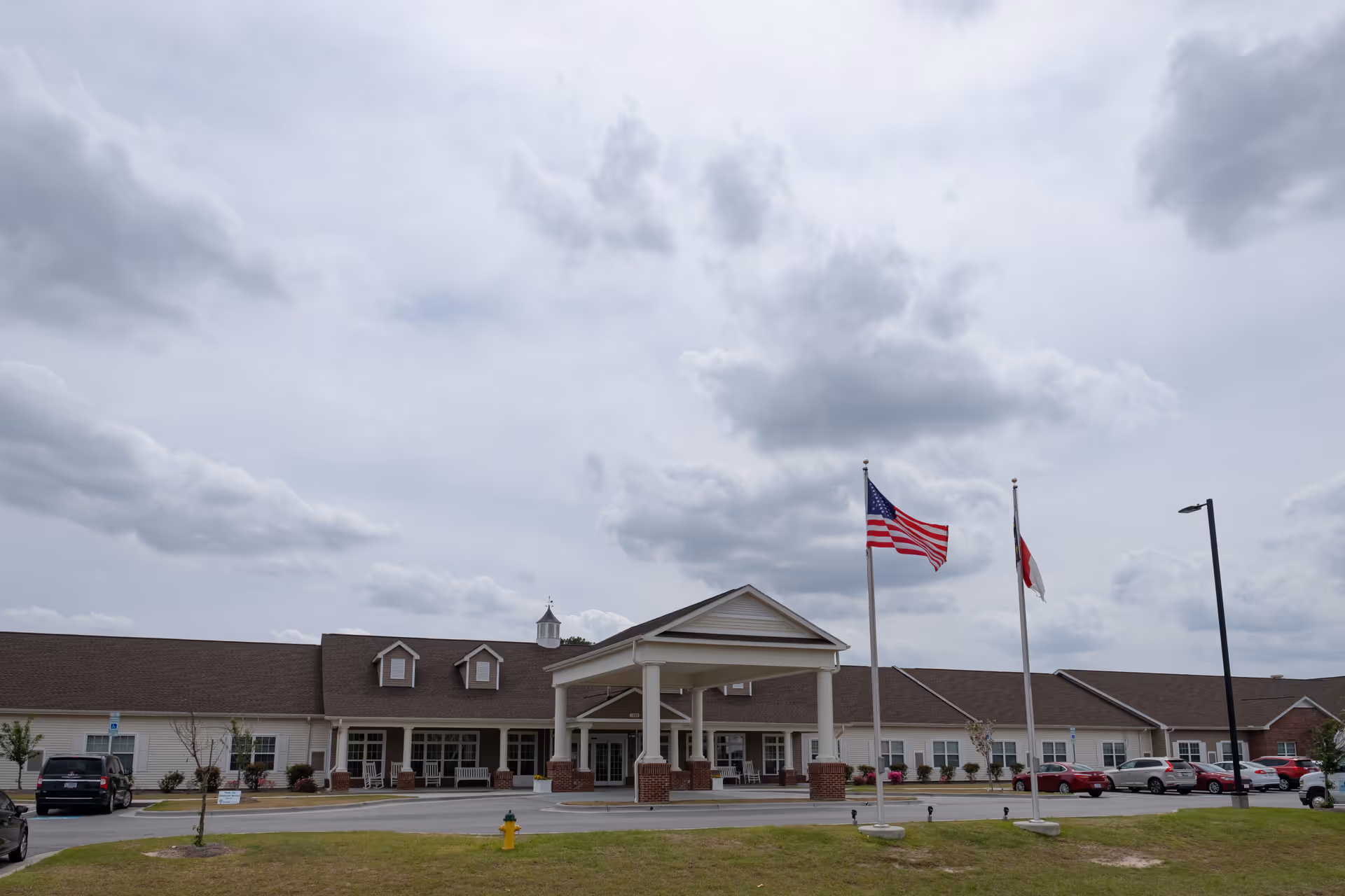 Front exterior of a single-story senior living building with a covered porte-cochère, American flag, and parking area.