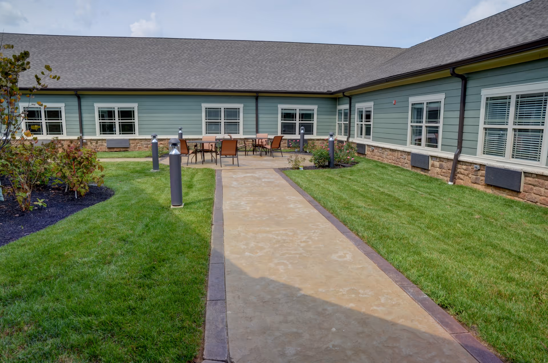 Outdoor courtyard area with a paved walkway leading to a patio with tables and chairs, surrounded by green grass and plants, adjacent to a single-story building with multiple windows and a gray roof under a partly cloudy sky.