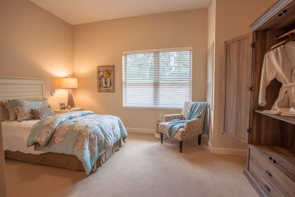 A cozy bedroom with a bed covered in a floral patterned comforter and multiple pillows. A nightstand with a lamp, clock, and picture frame is beside the bed. There is a window with blinds letting in natural light, an upholstered armchair with a blue throw blanket, and an open wooden wardrobe with a white robe hanging inside.