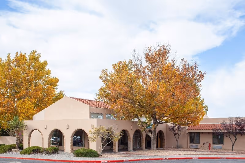 Exterior view of Las Palomas Center building with beige stucco walls, arched windows, and a red tile roof. Trees with autumn-colored leaves surround the building under a partly cloudy sky.
